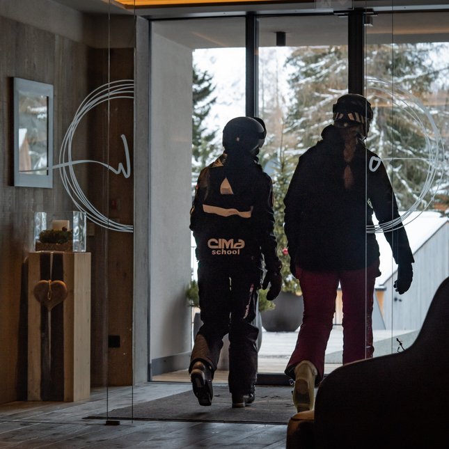 Il tuo hotel in Val Pusteria: Olangerhof Due persone in abbigliamento da sci escono da un edificio verso un paesaggio innevato.